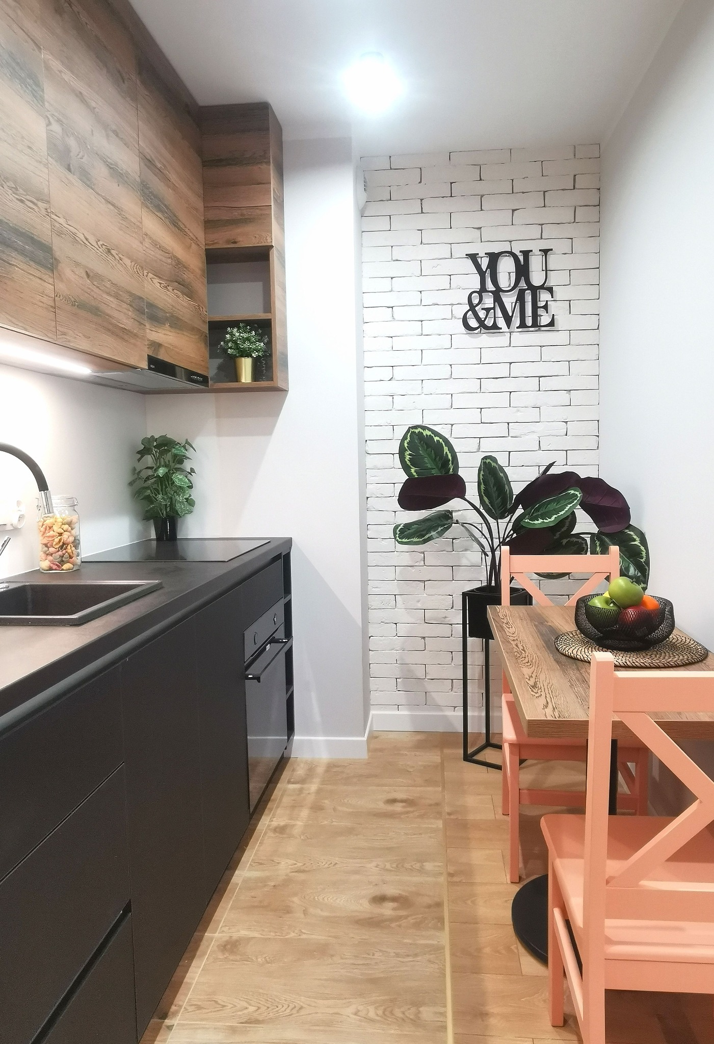 Nowoczesna kuchnia z drewnianymi szafkami i blatem w kolorze antracytu, biała ściana z cegły dekoracyjnej z napisem 'YOU & ME', drewniany stół i krzesła w odcieniu łososiowym, rośliny doniczkowe.