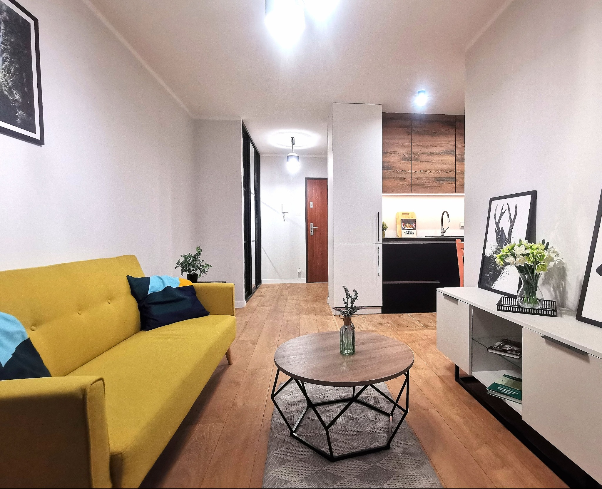 Jasne wnętrze mieszkania z żółtą sofą, okrągłym stolikiem kawowym i białą szafką RTV z dekoracjami, widok na otwartą kuchnię z drewnianymi akcentami.