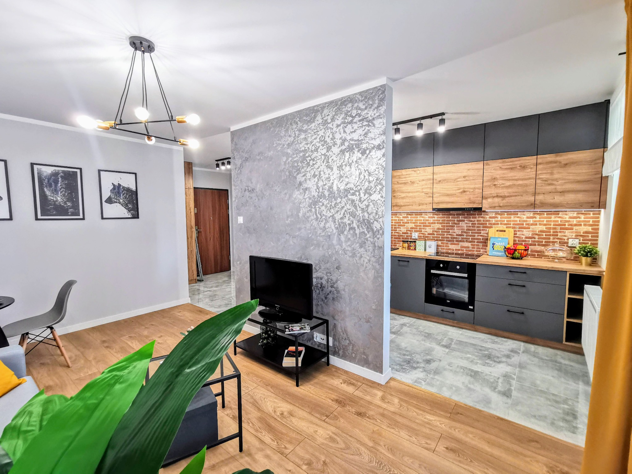 Nowoczesne wnętrze mieszkania z drewnianą podłogą, szarą ścianą z efektem dekoracyjnym, czarnym stolikiem pod telewizor i otwartą kuchnią z szarymi szafkami i ceglaną ścianą nad blatem.