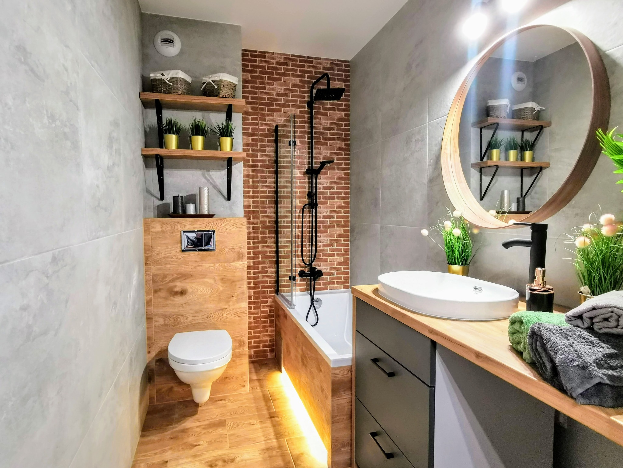 Nowoczesna łazienka z szarą płytką imitującą beton, ceglaną ścianą prysznicową, drewnianymi akcentami i oświetleniem LED pod wanną, uzupełniona okrągłym lustrem i dekoracyjnymi roślinami w złotych...
