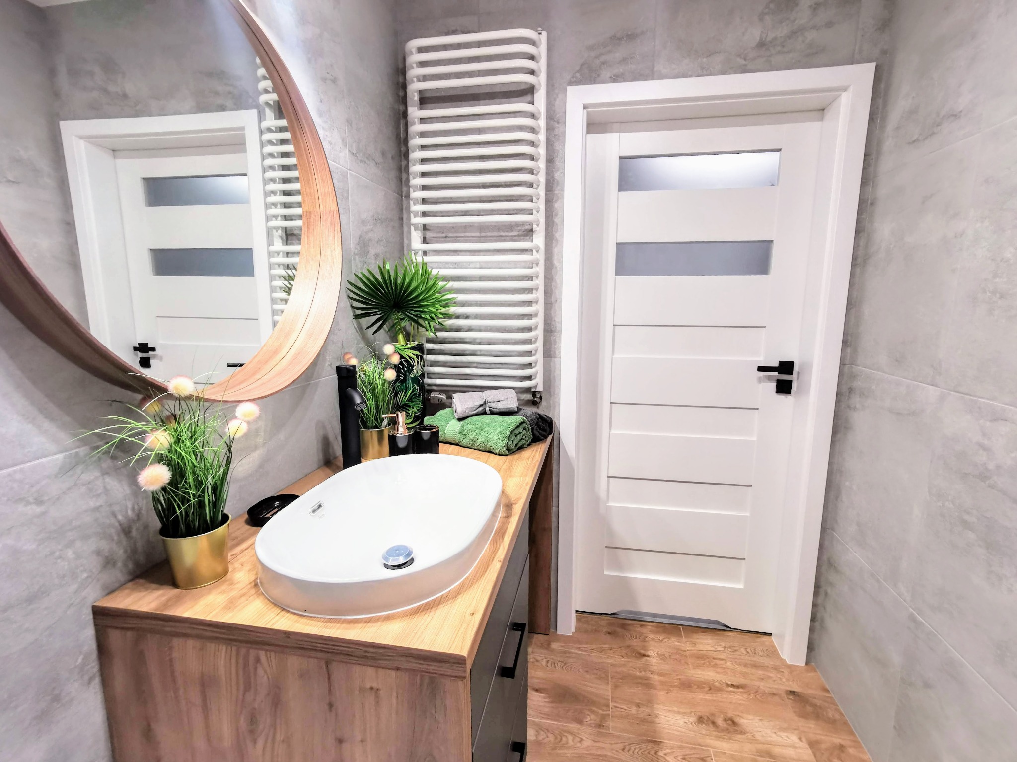 Nowoczesna łazienka z owalnym lustrem, umywalką nablatową na drewnianym blacie, białym grzejnikiem drabinkowym i drzwiami z przeszkleniami, wykończona szarymi płytkami imitującymi beton.