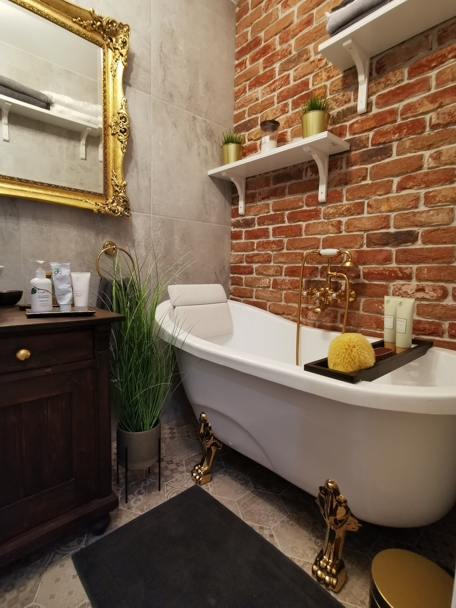 Retro łazienka z wolnostojącą wanną na złotych nóżkach, ceglaną ścianą, lustrem w złotej ramie i drewnianą szafką z kosmetykami.