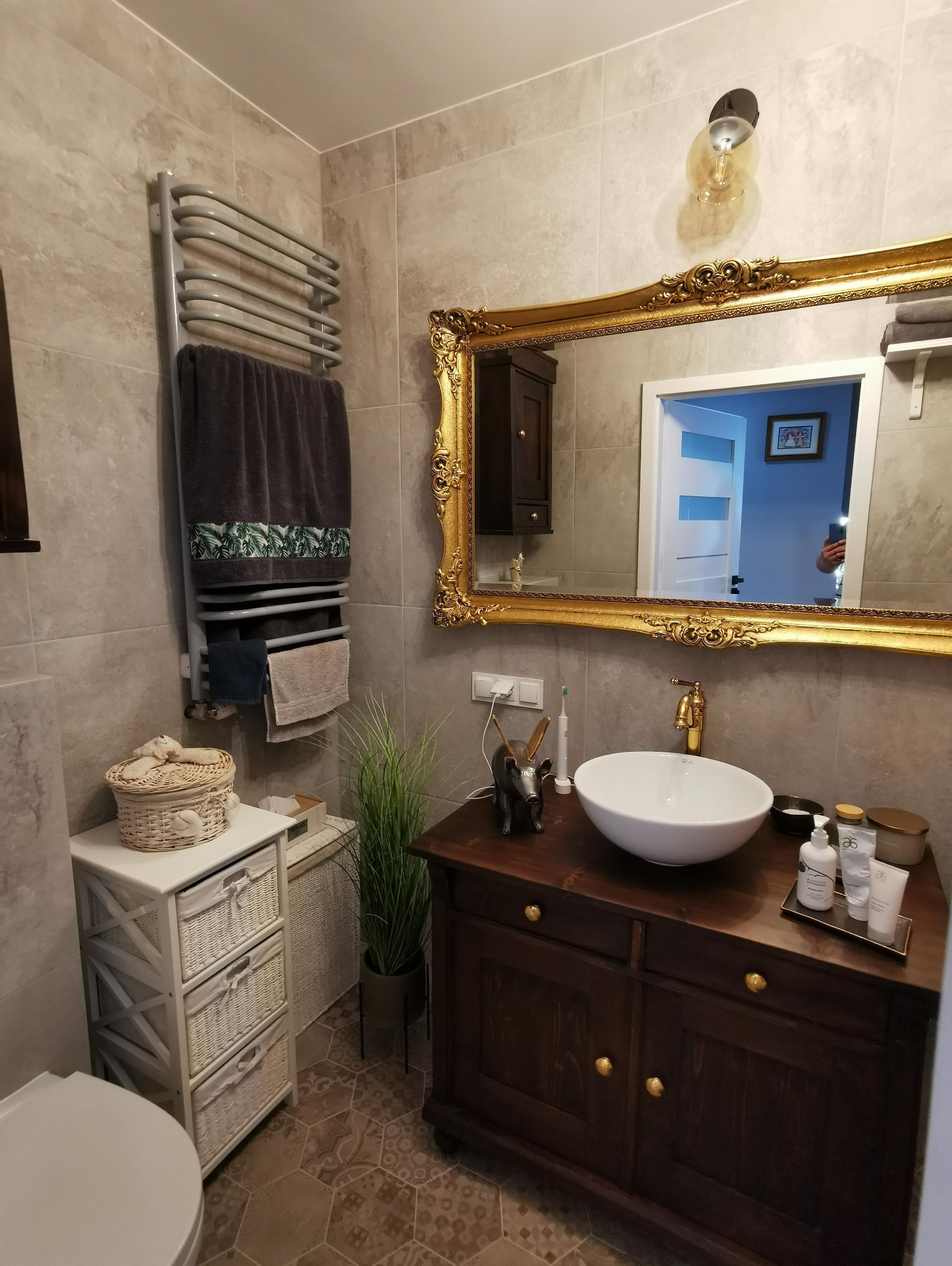 Umywalka nablatowa w okrągłej misie, złota bateria, ciemna szafka pod umywalką z ozdobnymi gałkami, duże lustro w bogato zdobionej złotej ramie, grzejnik drabinkowy z ręcznikami w łazience...