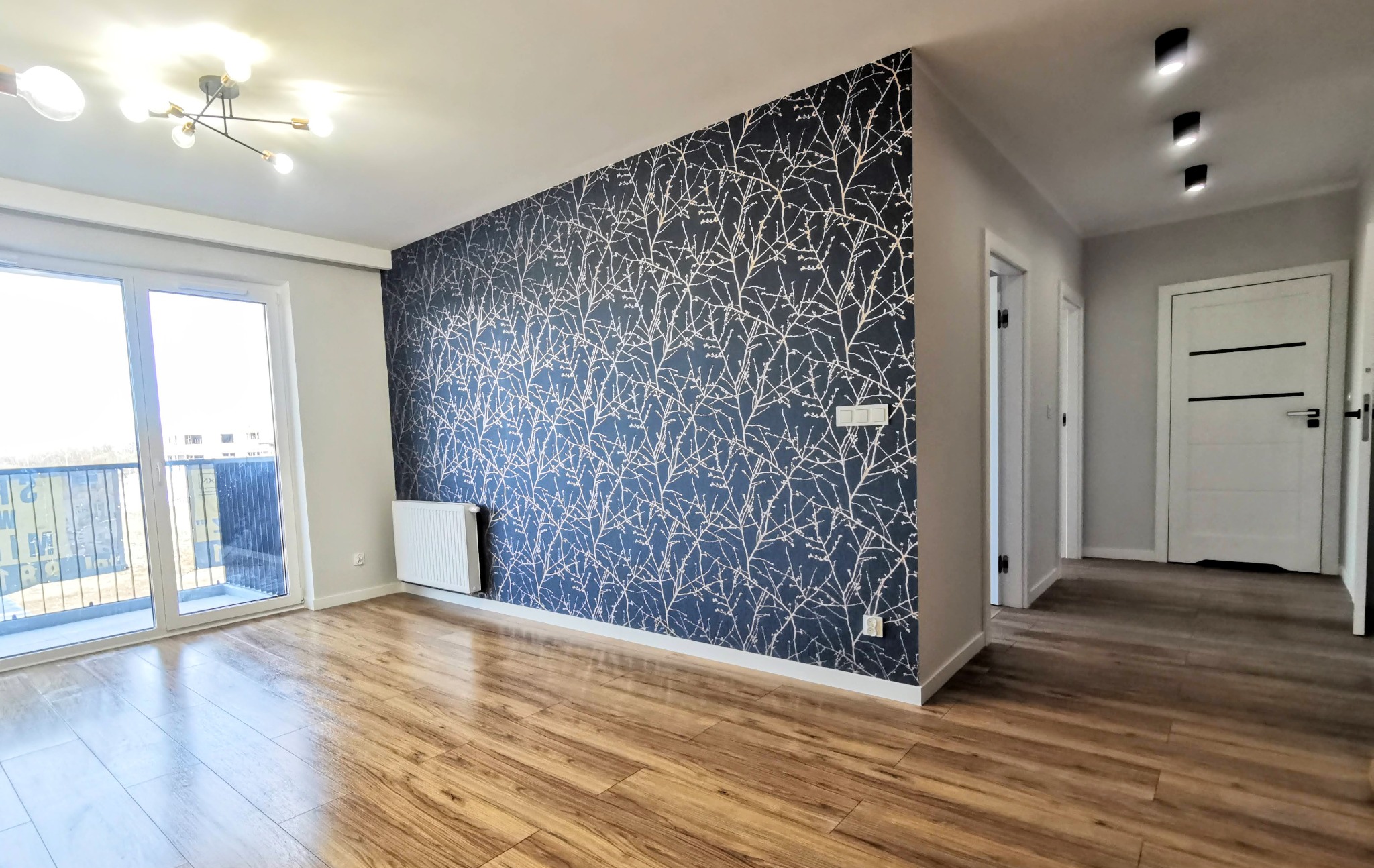 Pusty pokój z drewnianą podłogą, ścianą z tapetą w motywy gałązek i widokiem na balkon przez przesuwne drzwi, oświetlony nowoczesną lampą sufitową.