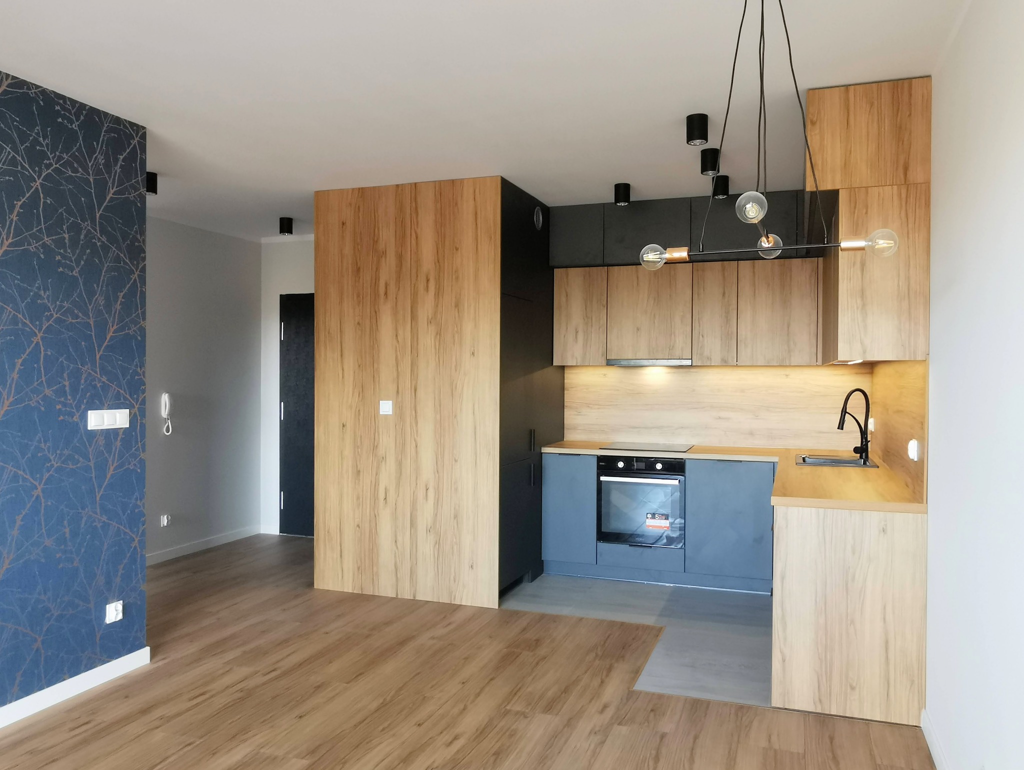 Nowoczesna kuchnia z drewnianymi frontami szafek, szarymi elementami i designerską lampą wiszącą, widok na fragment salonu z tapetą w gałązki.