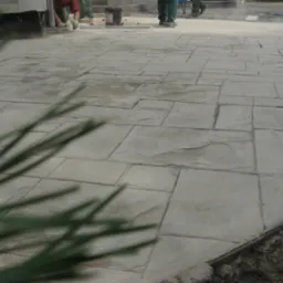 Szara, stemplowana nawierzchnia imitująca kamień, z widocznymi pracownikami w tle i rozmazanym fragmentem rośliny w pierwszym planie.