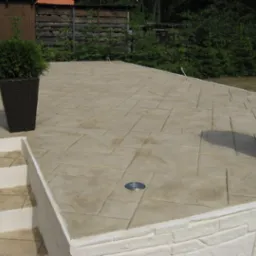 Stylowy taras z betonu stemplowanego imitującego kamień, z oświetleniem punktowym w podłodze, otoczony zielenią i dekoracyjnymi donicami z roślinami.