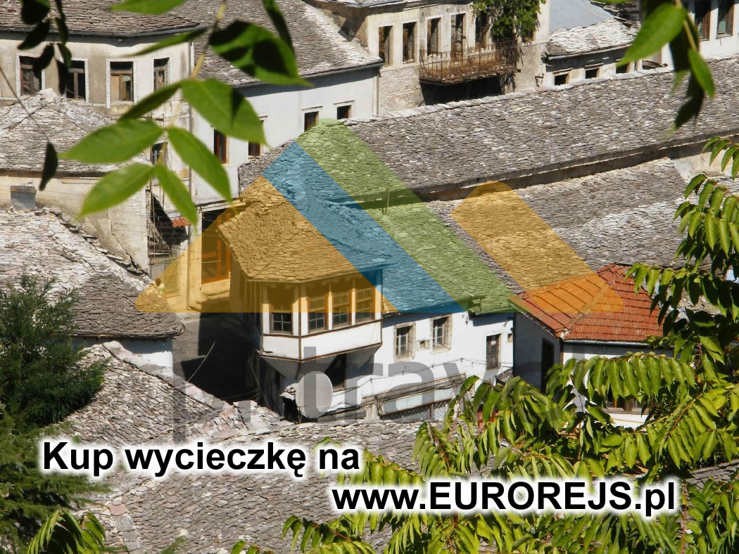 Widok z góry na starą zabudowę miejską z dachami pokrytymi szarymi płytkami, z widoczną zielenią drzew w kadrze, na pierwszym planie liście, w rogu widoczny adres strony internetowej.