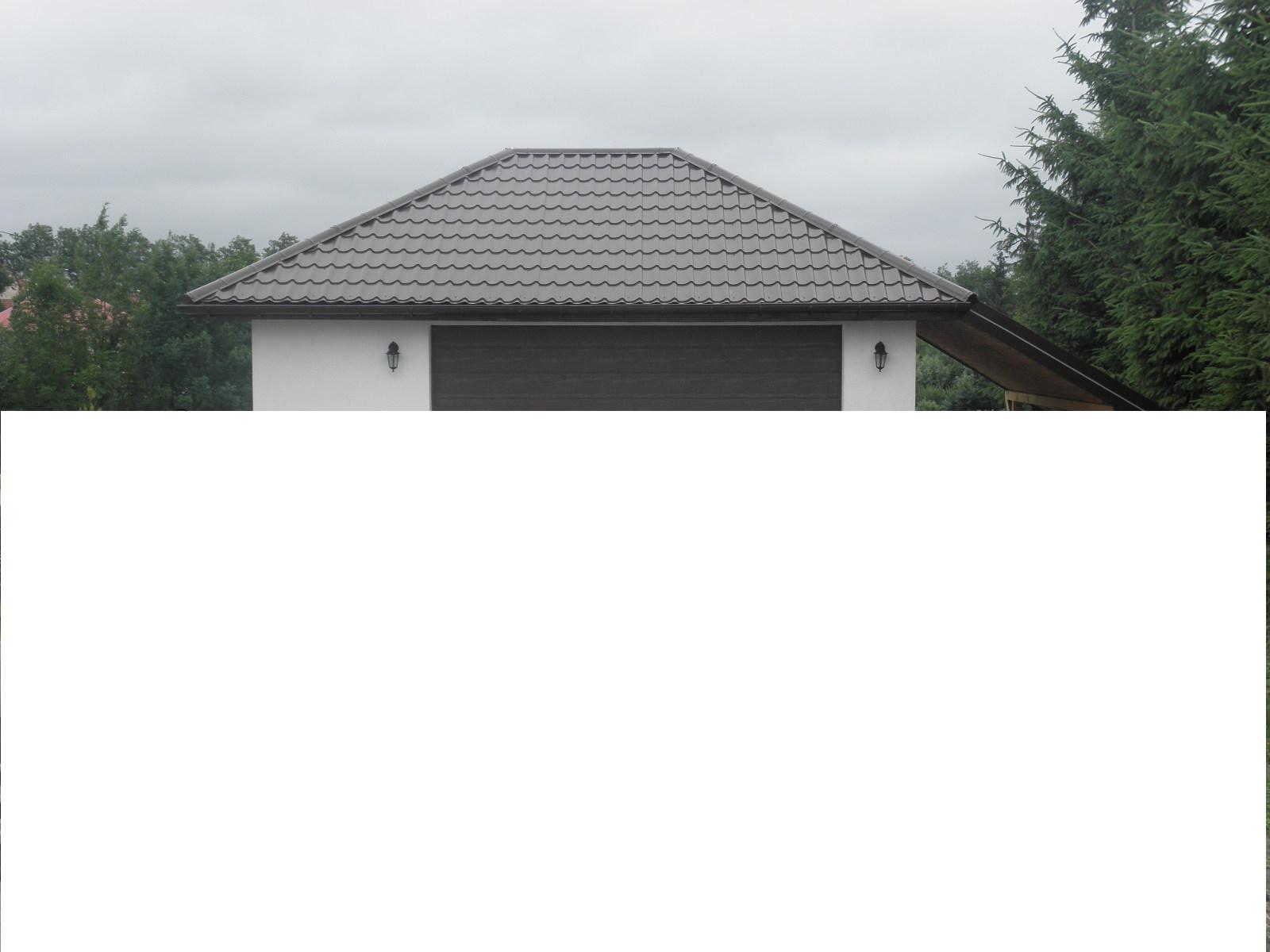 Szary dach z dachówki ceramicznej na białym garażu z brązową bramą, oświetlony dwoma lampami ściennymi, na tle pochmurnego nieba i zielonych drzew.