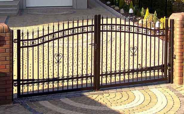 Dwuskrzydłowa, metalowa brama wjazdowa z kutymi zdobieniami w kształcie serc, osadzona między ceglanymi słupkami na posesji z brukowanym podjazdem.