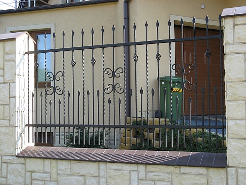 Ciemne, metalowe ogrodzenie z ozdobnymi elementami w kształcie motyli i grotów, na niskiej podmurówce z jasnego kamienia, z zielonym pojemnikiem na śmieci i brązową bramą garażową w tle.